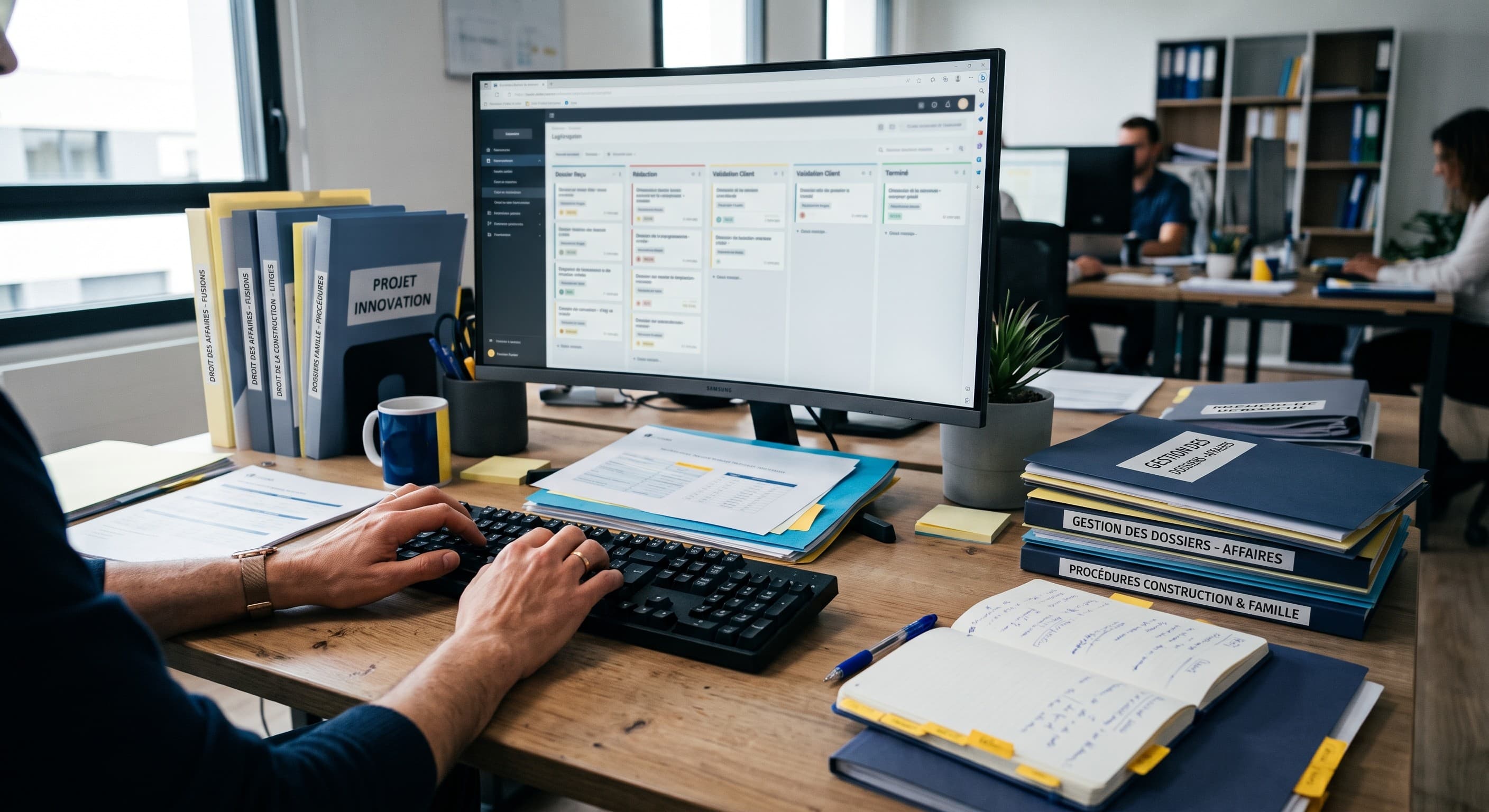 Un poste de travail dans un cabinet d'avocats : des mains sur un clavier, un écran affichant un suivi de dossiers, des classeurs de droit des affaires et de procédures, un carnet de notes annoté.